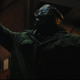 The Riddler, wearing a plastic mask over his face and glasses, unspools a roll of duct tape.